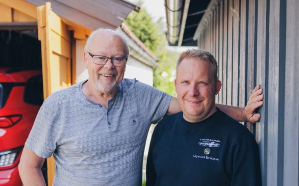 Elbileier Dag Røneid og elektriker Anders Bakke står smilende side om side foran garasje med rød elbil parkert